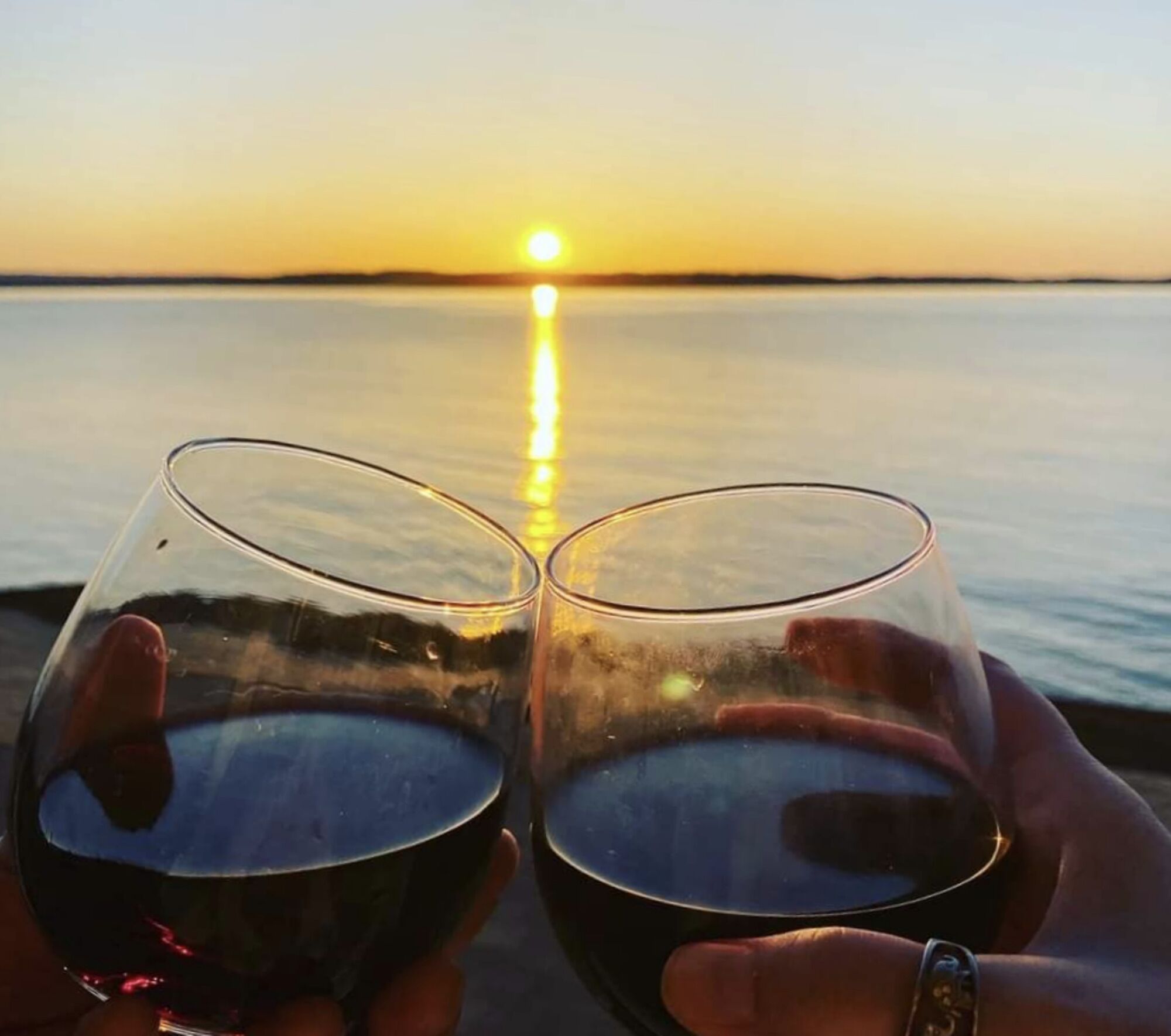 Dos copas de vino tinto sostenidas por manos al atardecer sobre el agua.