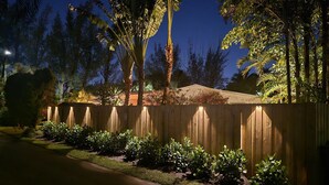 A wooden fence with a garden in front, surrounded by various trees and plants.