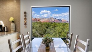 Uma área de refeições com mesa e cadeiras, vista para formações rochosas avermelhadas e uma parede de tijolos.