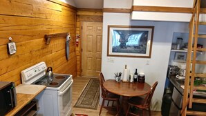 A compact kitchen with a stove, microwave, and sink. There is a dining table with chairs and a rug with apple designs. A framed picture hangs on the wall.