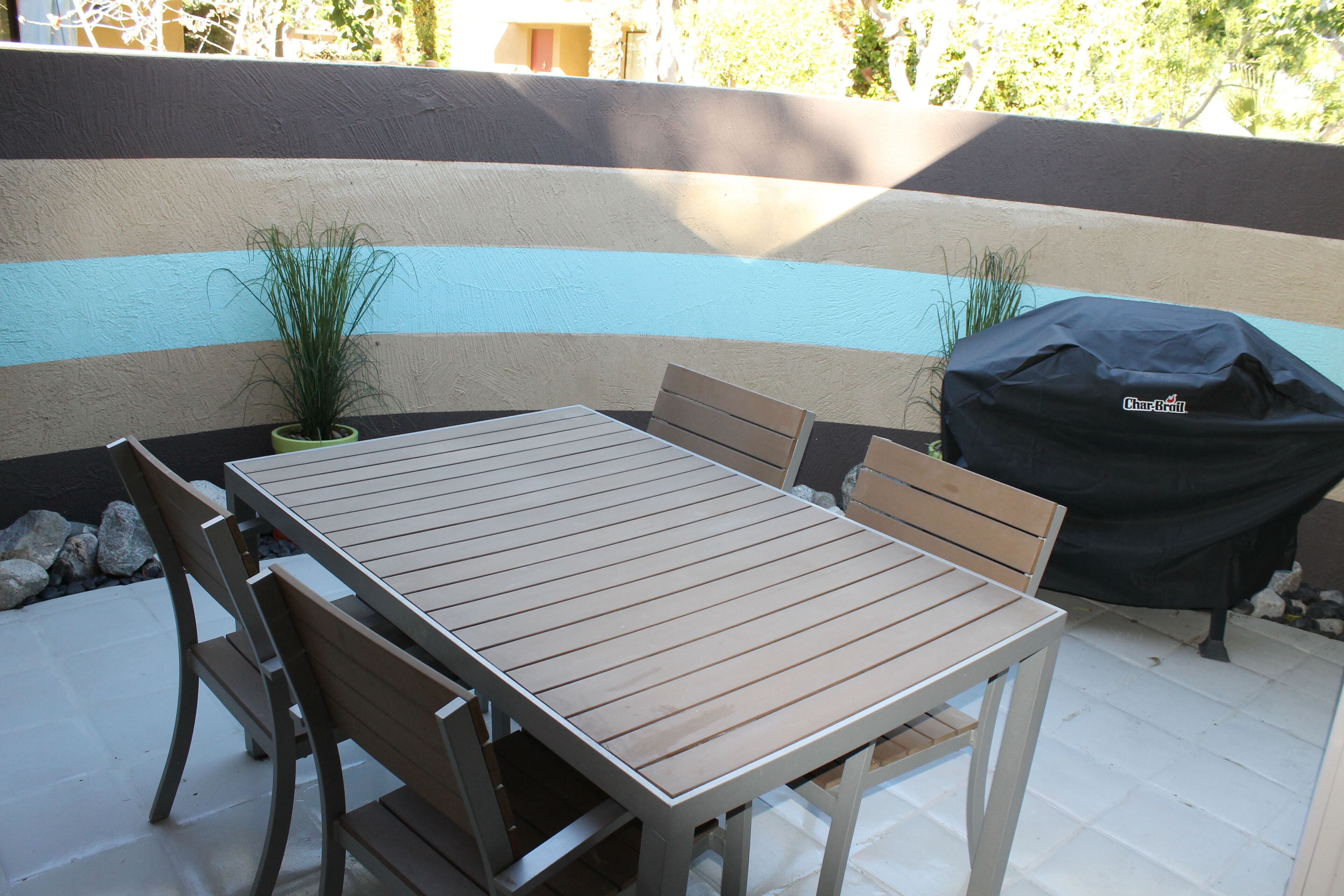 An outdoor patio with a wooden table and chairs, a barbecue grill, and a potted plant.