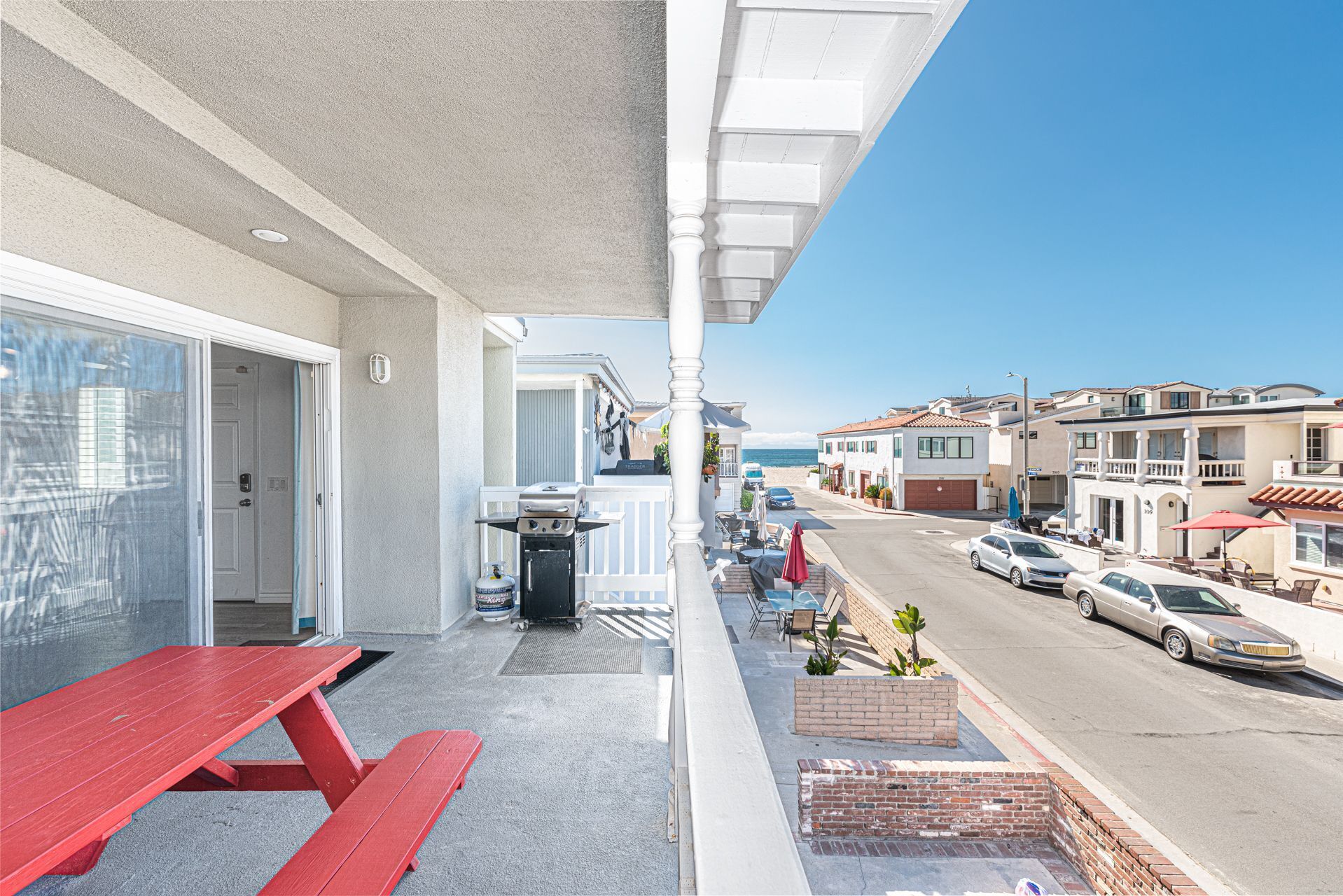 A balcony with a red picnic table, a barbecue grill, and a view of a street with cars and buildings.