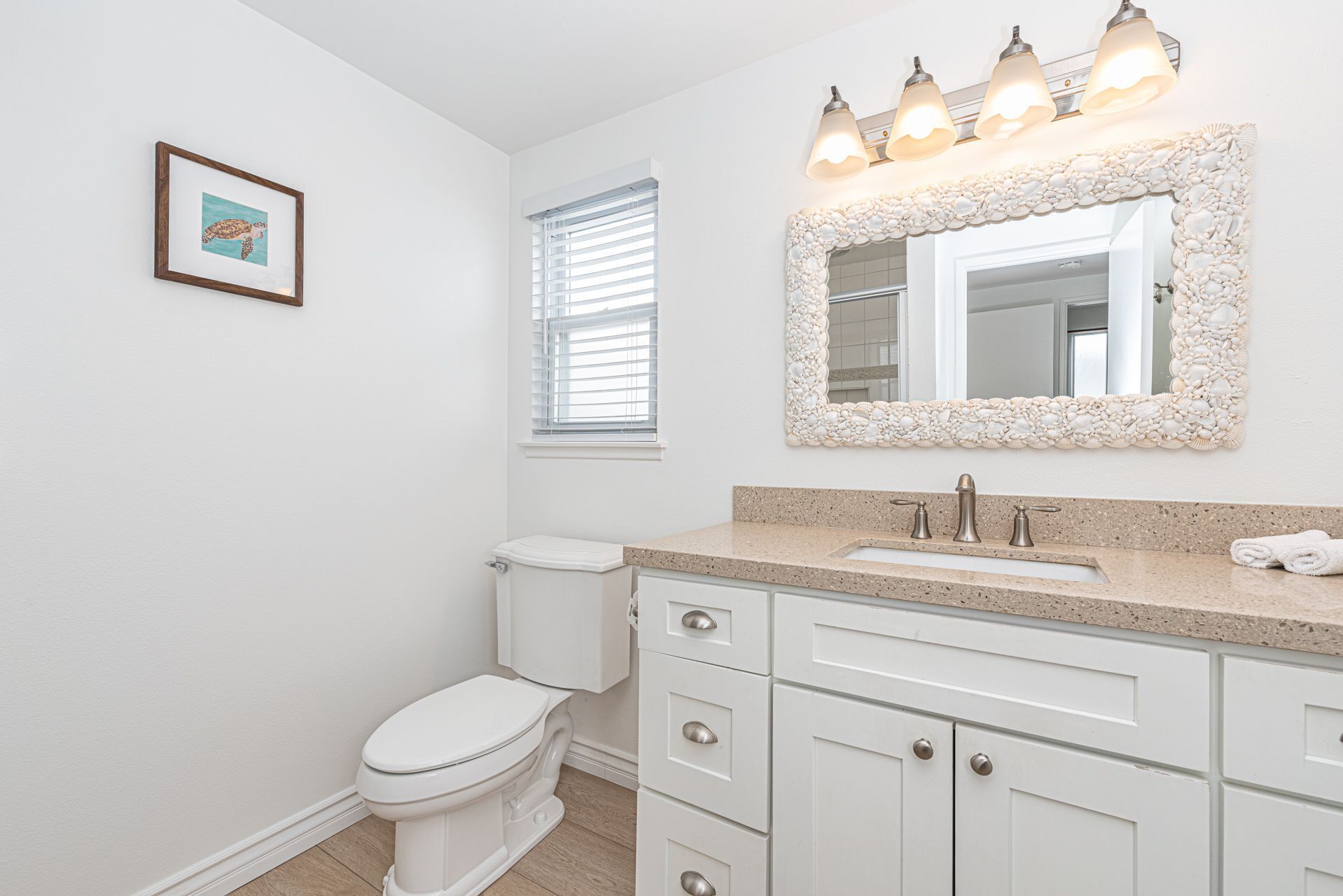 Une salle de bain équipée de toilettes, d’un lavabo et d’un meuble-lavabo avec des tiroirs. Un tableau encadré est accroché au mur, et il y a une fenêtre avec des stores.