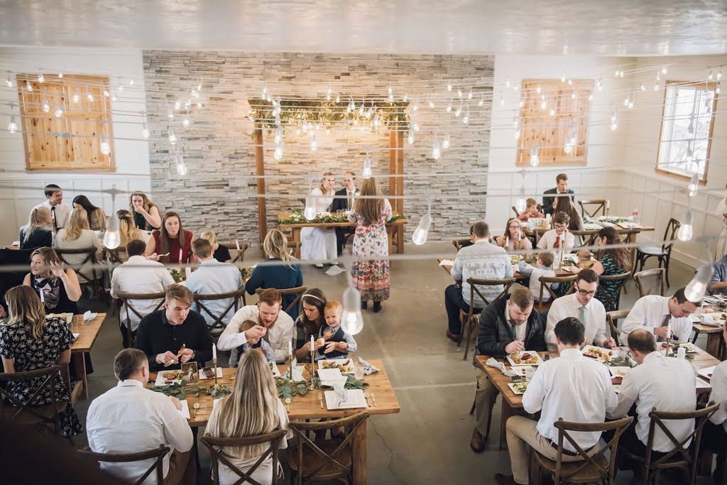 A dining area with tables arranged in a semi-circle, a stone wall in the background, and overhead string lights.