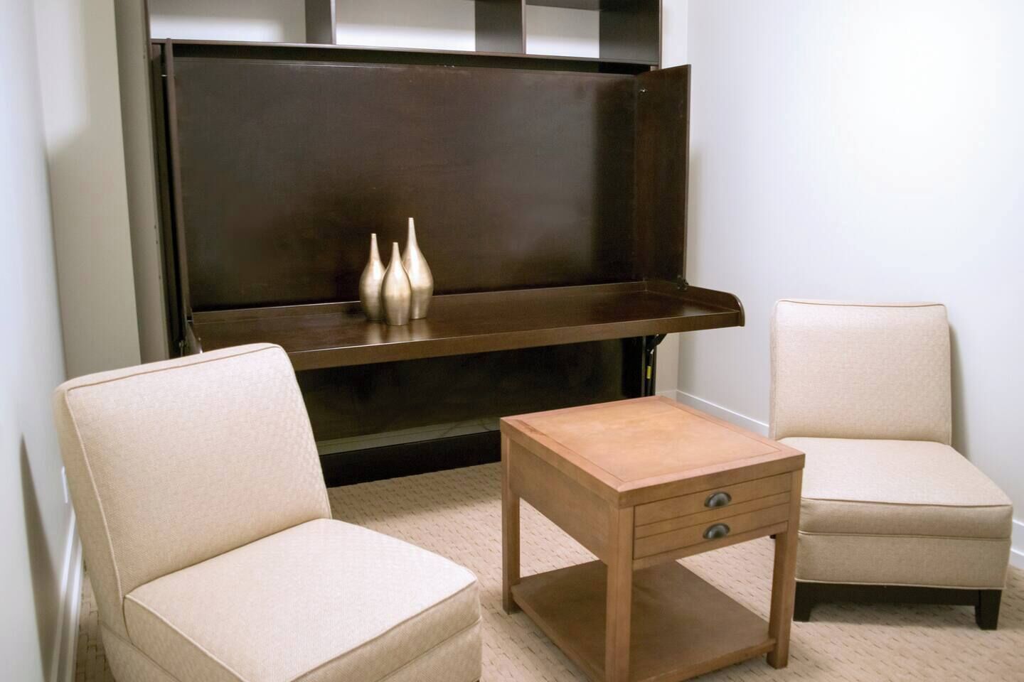 Een kamer met twee beige fauteuils, een klein houten tafeltje met een lade, en een donker houten kast met drie vazen erop.