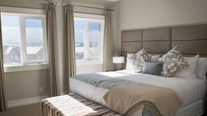 Quarto de hotel com uma cama grande, um banco, uma mesa de cabeceira com abajur e vista para edifícios pela janela.
