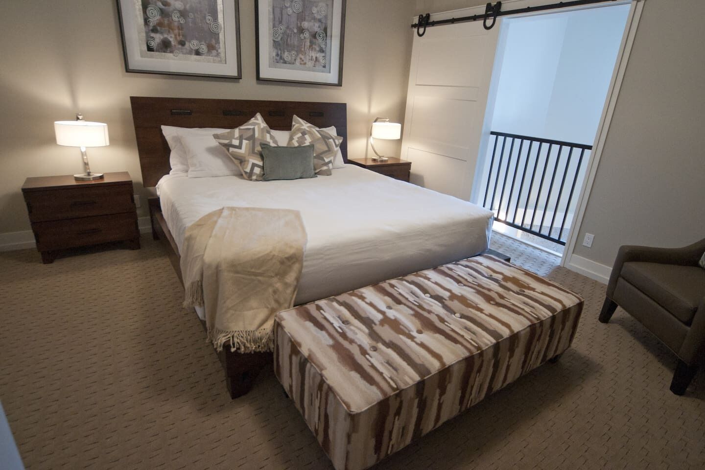 Une chambre avec un grand lit, deux tables de chevet avec des lampes, un banc, un balcon avec une balustrade et deux œuvres d’art encadrées accrochées au mur.