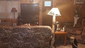A cozy living room with a patterned sofa, a television, a lamp with a forest scene, and a wooden coffee table. - Horsehead Lake Retreat (Big Discounts in June Call 414-378-7273)  (Presque Isle)