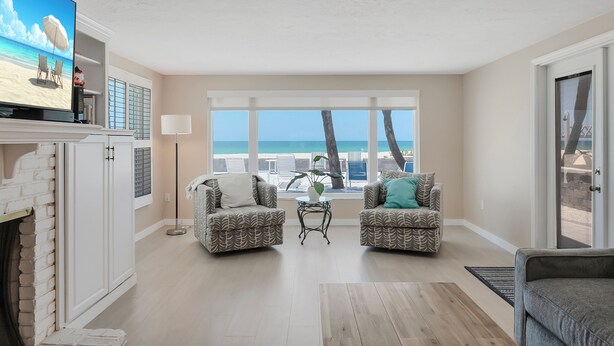 Ein Wohnzimmer mit Fernseher, der eine Strandansicht zeigt, zwei gemusterten Sesseln, einem kleinen Tisch mit einer Pflanze und Meerblick durch große Fenster.