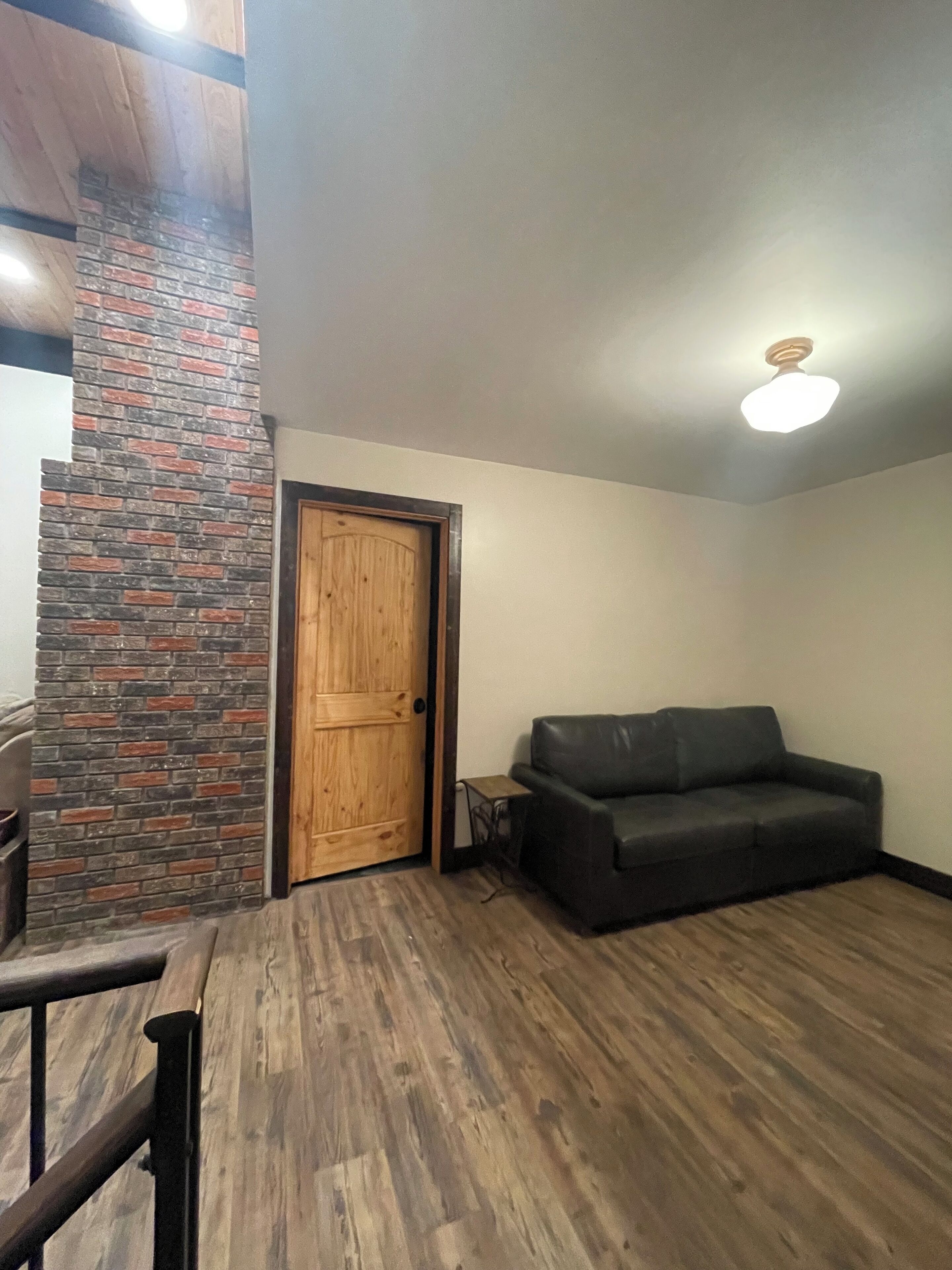 Une pièce avec une porte en bois, un mur en briques apparentes, un canapé en cuir noir et un plancher en bois.
