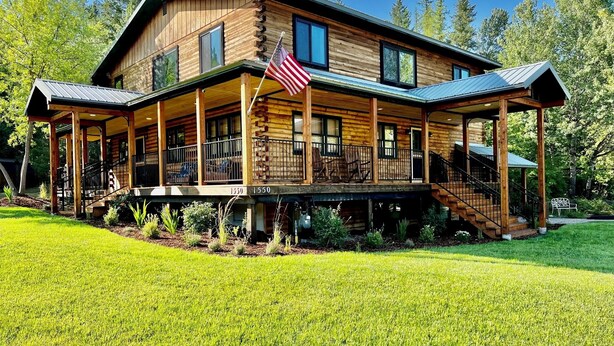 A two-story log cabin with a covered porch, a flagpole, and a staircase leading to the upper level. - Cozy log home sleeps 14 in beds 5.5 baths near Glacier Park and Whitefish Resort (Columbia Falls)
