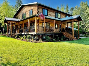 A two-story log cabin with a covered porch, a flagpole, and a staircase leading to the upper level.