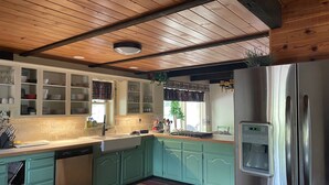 A kitchen with wooden ceiling beams, teal cabinets, a stainless steel refrigerator, and a window with a view of greenery outside. - Cozy log home sleeps 14 in beds 5.5 baths near Glacier Park and Whitefish Resort (Columbia Falls)