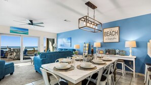 A dining area with a white table set for six, blue sofas in the background, and a view of the ocean through large windows. - Touch of Paradise-Gulf Front Home Newly Renovated. Pup Friendly. Sleeps 12 (Navarre)