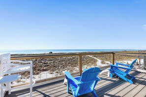 Una terraza con dos sillas Adirondack azules y una silla blanca, con vista a una playa y dunas.