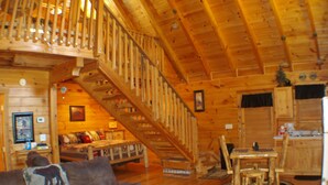 A wooden cabin interior with a staircase, a living room with a couch, a dining area, and a kitchen.