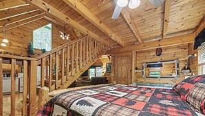 A log cabin interior with a bed, staircase, kitchen, and a large window.