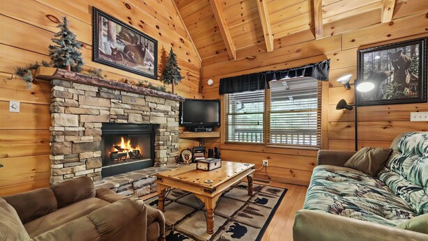 A log cabin interior with a stone fireplace, wooden furniture, and framed pictures on the walls.