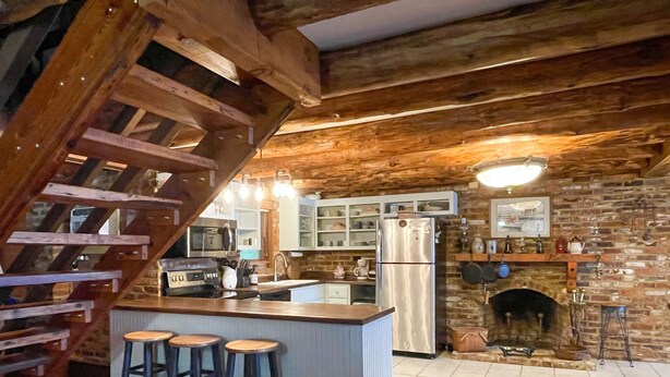 A rustic kitchen with a wooden staircase, a central island with stools, a refrigerator, and a fireplace.