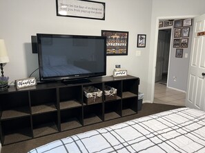Quarto com uma grande TV de tela plana, uma cama com edredom xadrez, uma pequena mesa de cabeceira com abajur, e uma parede com fotos emolduradas e uma frase motivacional.