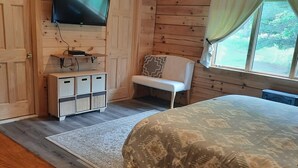 A wooden cabin room with a bed, a TV mounted on the wall, a bench, and a window with curtains.