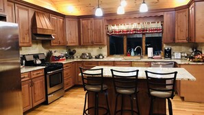 A kitchen with wooden cabinets, a stainless steel refrigerator, a stove, and a sink. There are three bar stools and a window with a curtain.