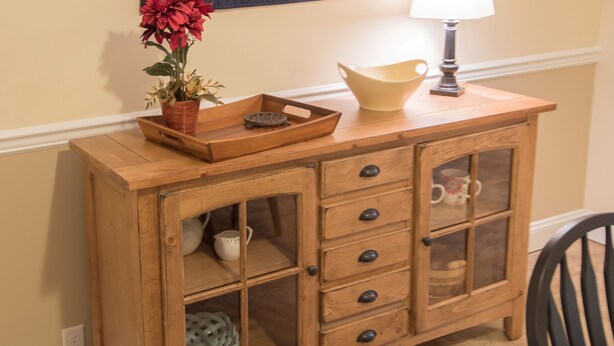 Un meuble en bois avec des tiroirs et un autre meuble avec une porte en verre, une courtepointe accrochée au mur, une plante en pot et une lampe de table.
