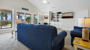 A spacious living room with a large blue sofa, a ceiling fan, and a view of the outdoors through large windows. - 4 Bedrooms, 4  Baths; Just Steps to the Ocean!  Fronts on Point St. (Edisto Beach)