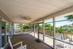 A covered outdoor deck with wooden flooring, white rocking chairs, and a hammock.