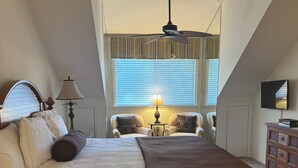 Een slaapkamer met een bed, twee stoelen, een lamp, een plafondventilator, een televisie en een raam met zonwering.