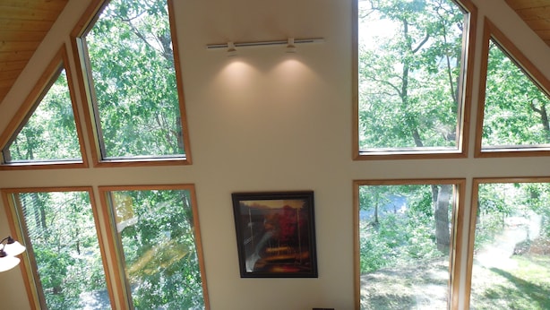 Een kamer met grote ramen met uitzicht op een bos. Er hangt een ingelijst beeld aan de muur en er staat een lamp op een tafel.