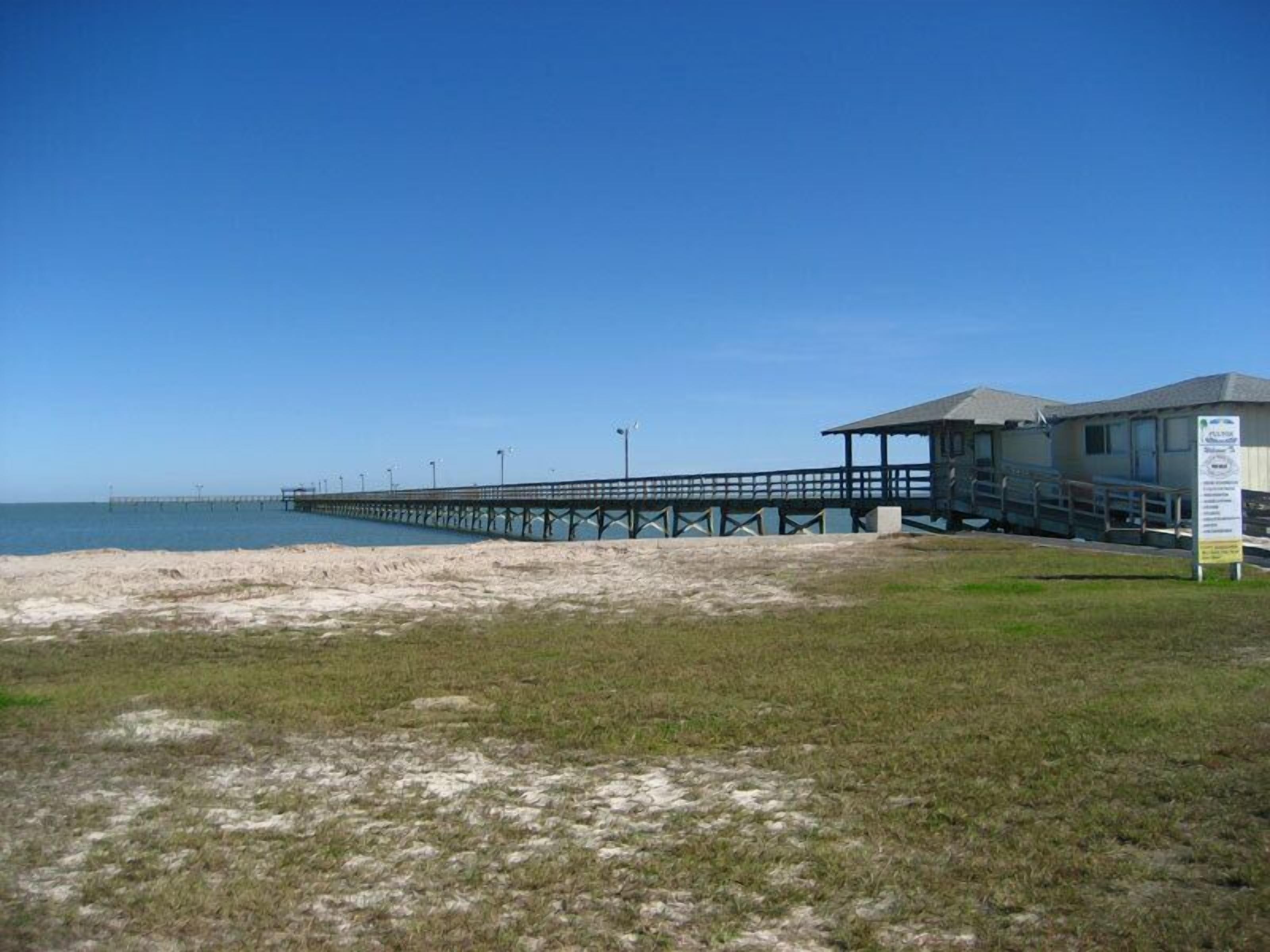 A beach with a pier extending into the water, a small building with a covered area, and a grassy area in the foreground.