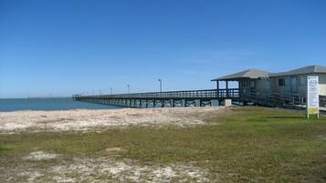 A beach with a pier extending into the water, a small building with a covered area, and a grassy area in the foreground.
