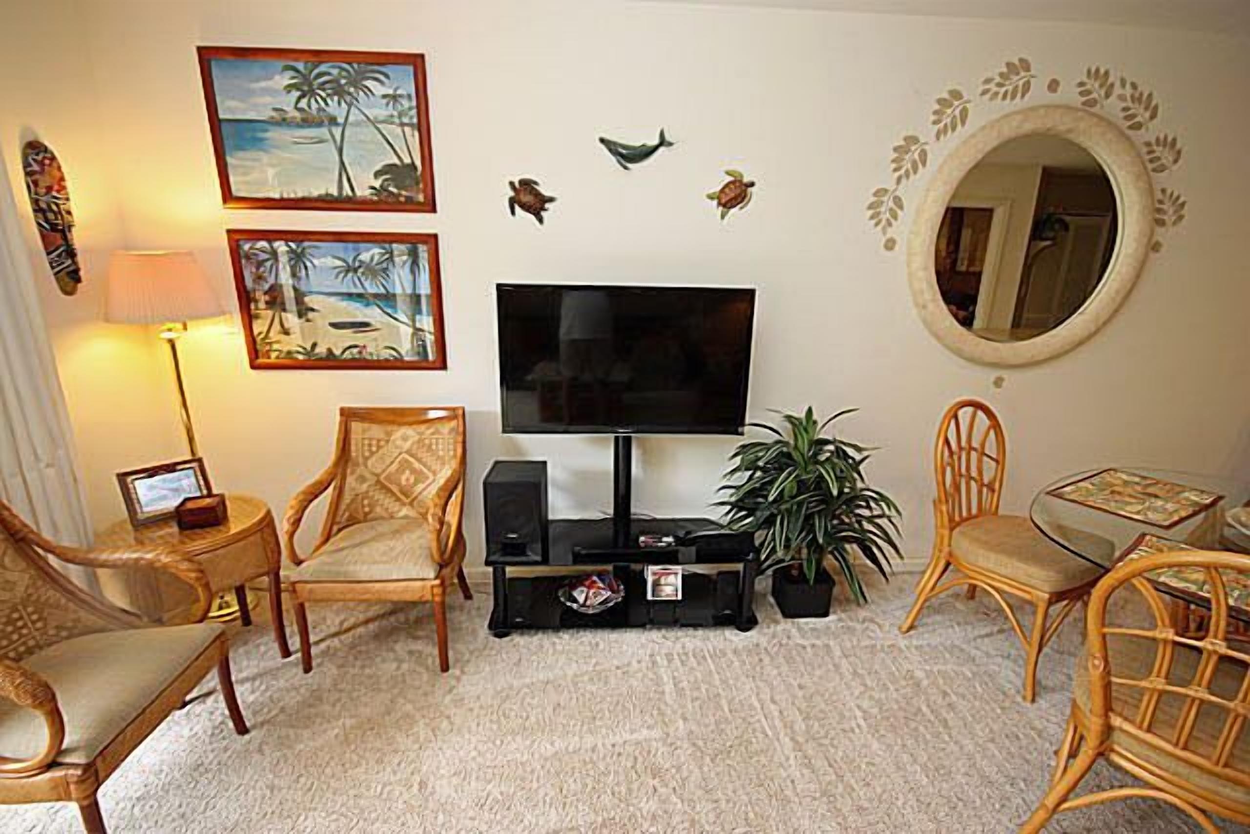 A living room with a flat-screen TV, two wooden chairs, a small table, a lamp, and wall decorations including a mirror and framed pictures.