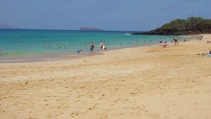 Una playa de arena con agua turquesa cristalina, un promontorio rocoso a lo lejos y gente disfrutando de la playa.