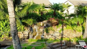 Un jardín tropical con cascada, palmeras y una zona de piscina con sillones de descanso.