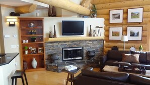 A cozy living room with a stone fireplace, a flat-screen TV mounted above it, a leather sofa, and a wooden bookshelf filled with decorative items.