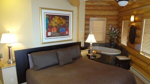 A log cabin-style room with a large bed, two bedside lamps, a framed artwork, a small table with a lamp, and a bathroom with a bathtub and window blinds.