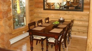 A wooden log cabin dining room with a large table, chairs, and a window with a view of trees.