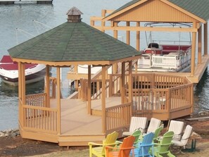 A wooden gazebo with a green roof and a deck, surrounded by chairs and a boat docked nearby.