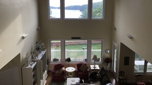 A spacious room with a high ceiling, large windows offering a view of a lake and hills, a cozy seating area with a red armchair and a small table, and a decorative shelf with glassware and figurines. - Luxury on Lake Keowee - FALL & Summer 2027 NOW OPEN (Seneca)