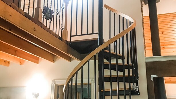 Escadaria em espiral com degraus de madeira e corrimãos pretos, que dá acesso a um andar superior.