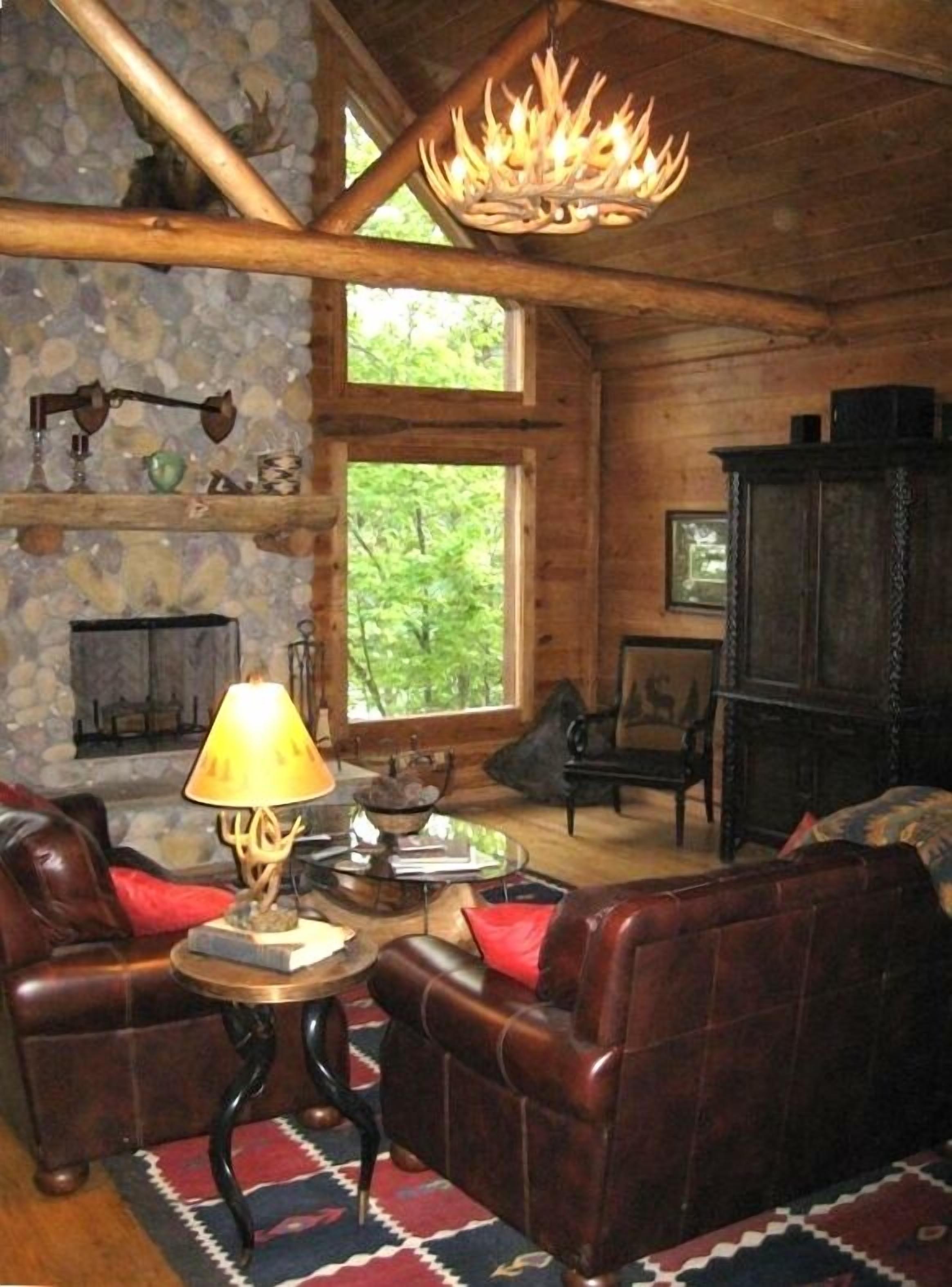 A rustic cabin interior with a stone fireplace, leather sofas, and a chandelier.