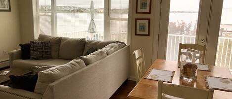 Ein Wohnzimmer mit einer Couch, einem Holzesstisch und Stühlen. Es gibt Fenster mit Jalousien, eine Tür und einen Blick ins Freie.