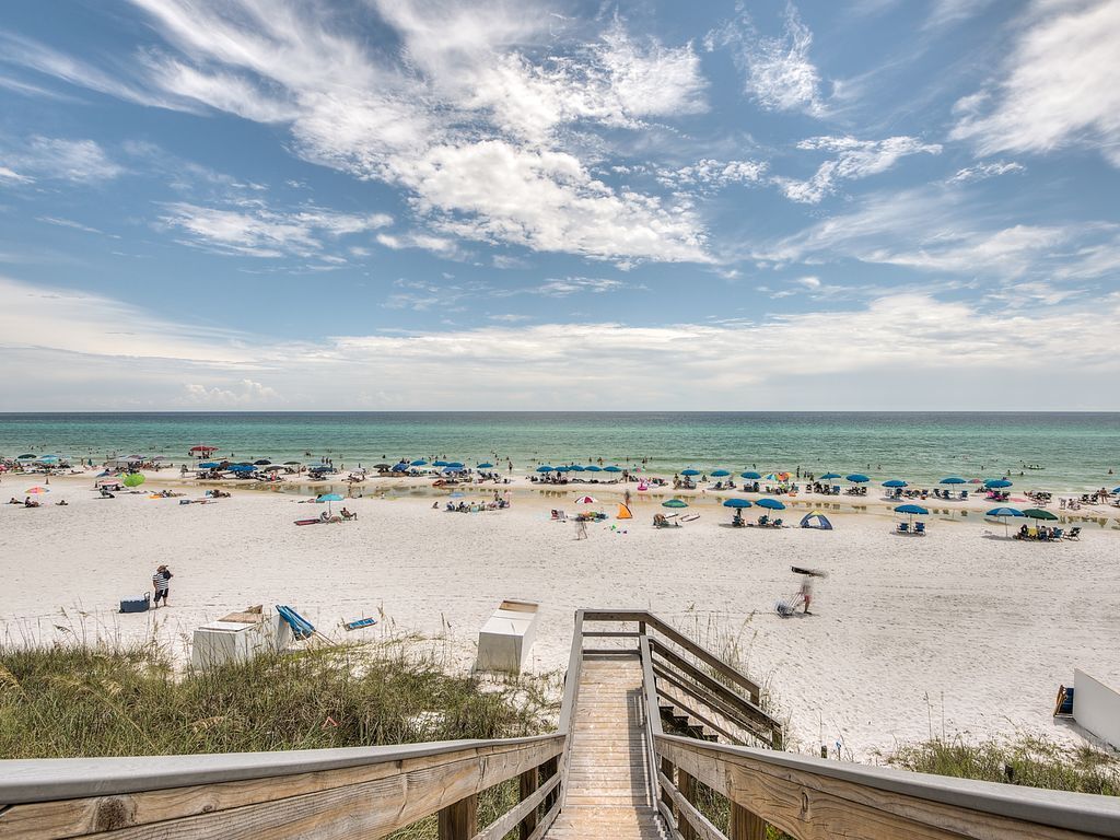 A beach with a wooden staircase leading down to the sand, umbrellas, and beachgoers.