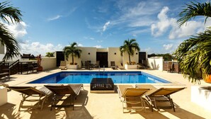 A swimming pool with lounge chairs and palm trees, surrounded by a white building with a flat roof.