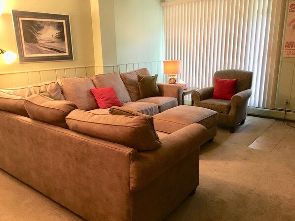 A living room with a brown sofa, a brown armchair, and a small table with a lamp. There is a framed picture on the wall and a window with blinds.