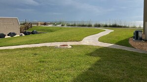 A grassy outdoor area with a concrete path, barbecue grills, and an air conditioning unit.