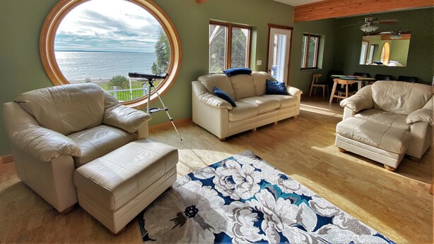 A living room with a large round window overlooking a body of water, a wooden ceiling, and a dining area with a table and chairs. - LaBarbe Bleu on Lake Michigan (Saint Ignace)