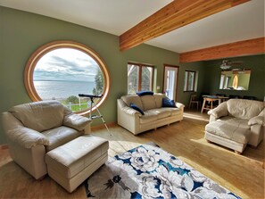 A living room with a large round window overlooking a body of water, a wooden ceiling, and a dining area with a table and chairs. - LaBarbe Bleu on Lake Michigan (Saint Ignace)